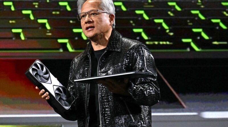 Male tech presenter on stage holding a large GPU in one hand and a slim laptop in the other, wearing a shiny black jacket with green-lit backdrop behind him.