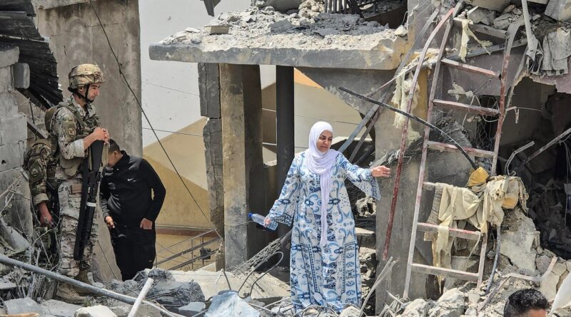 Woman in a white hijab and blue patterned dress stands amid rubble, gesturing, with two soldiers nearby at a destroyed building site.