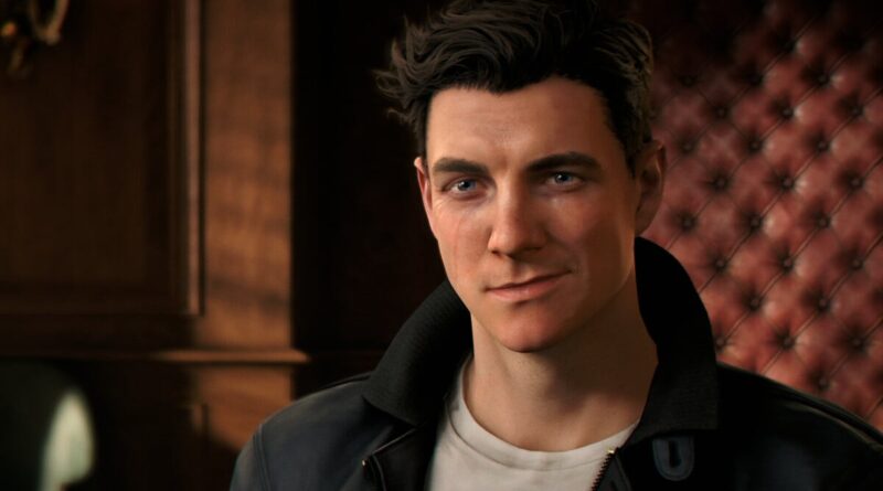 Close-up of a man with dark hair in a black leather jacket, smiling slightly, indoors with a red tufted wall in the background.