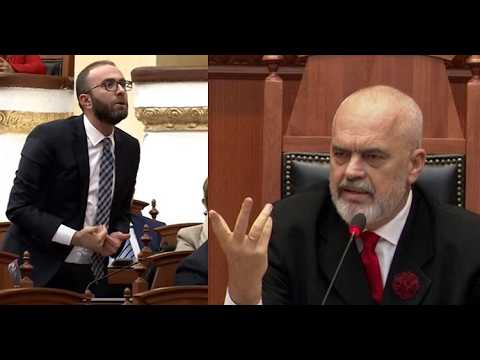 Split-screen of two men in a formal legislative chamber: left man in a suit with glasses gesturing, right man bald with a beard speaking at a microphone.