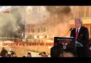 Live protest scene: man in a suit speaks at a podium as smoke rises and protesters gather outside a building; red on-screen text.