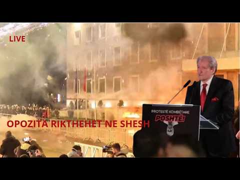 Live protest scene: man in a suit speaks at a podium as smoke rises and protesters gather outside a building; red on-screen text.