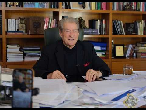 Older man in a dark suit sits at a cluttered desk in a study with shelves of books behind him, looking at the camera.