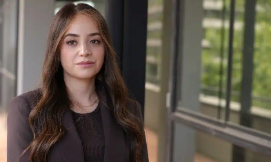Professional woman with long dark hair in a dark blazer standing outside a glass-front building near office windows.