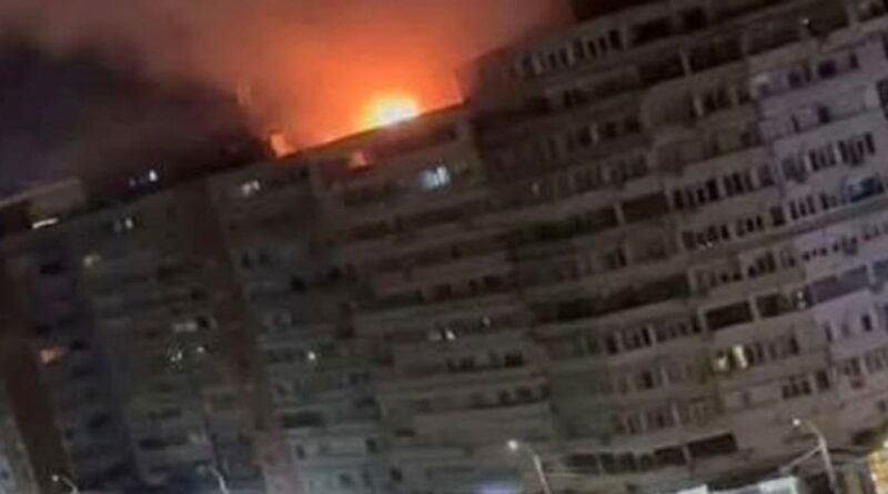 Nighttime scene of a large apartment building with a bright fire on the roof and smoke surrounding it.