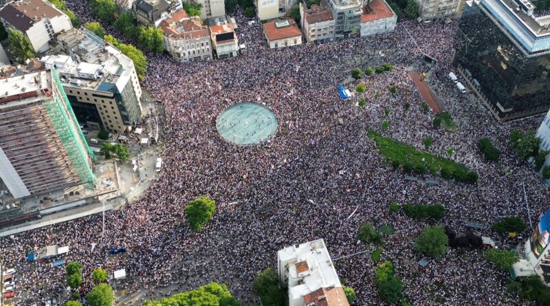 Aerial view of a dense crowd in a city square around a circular fountain, surrounded by buildings and trees.