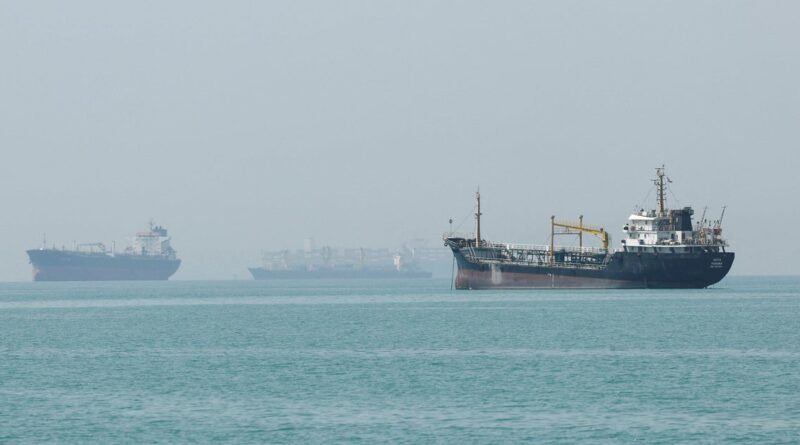 A large cargo ship sails on calm turquoise water with a hazy horizon and faint ships in the distance beneath a pale sky.