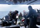Divers on a boat preparing dive gear: multiple scuba tanks, regulators, and wetsuits, with the open sea in the background.