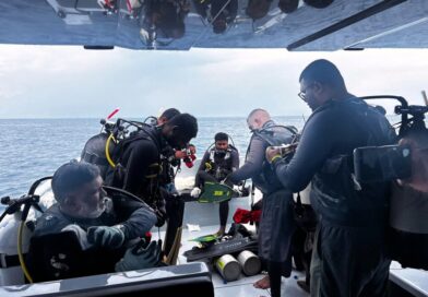Divers on a boat preparing dive gear: multiple scuba tanks, regulators, and wetsuits, with the open sea in the background.