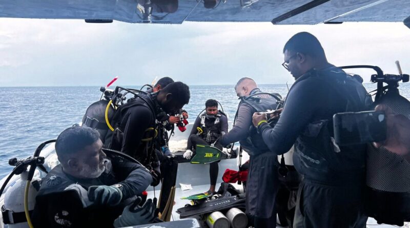 Divers on a boat preparing dive gear: multiple scuba tanks, regulators, and wetsuits, with the open sea in the background.