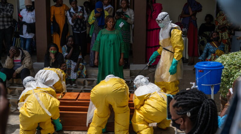 Mourners gather as people in yellow hazmat suits bury a coffin at a community burial. A healthcare worker in PPE oversees the process.