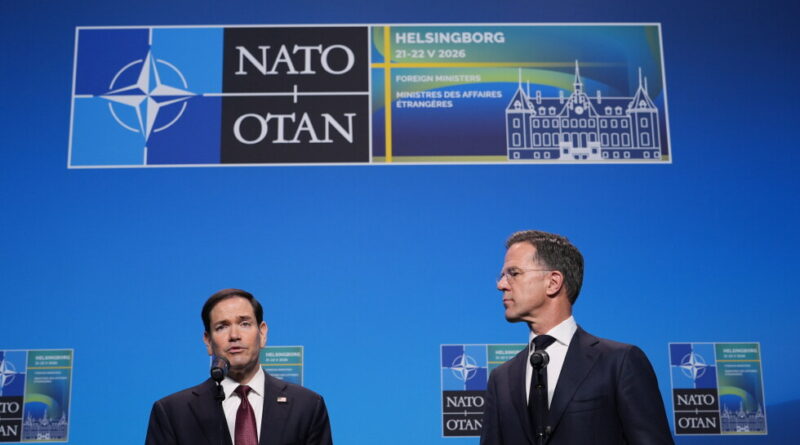 Two men in suits stand at podiums giving a press conference in front of a blue NATO backdrop with a large logo sign above them.