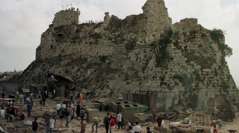 Stone fortress ruins atop a rocky hill, with debris and a crowd of people and vehicles gathered at the base.