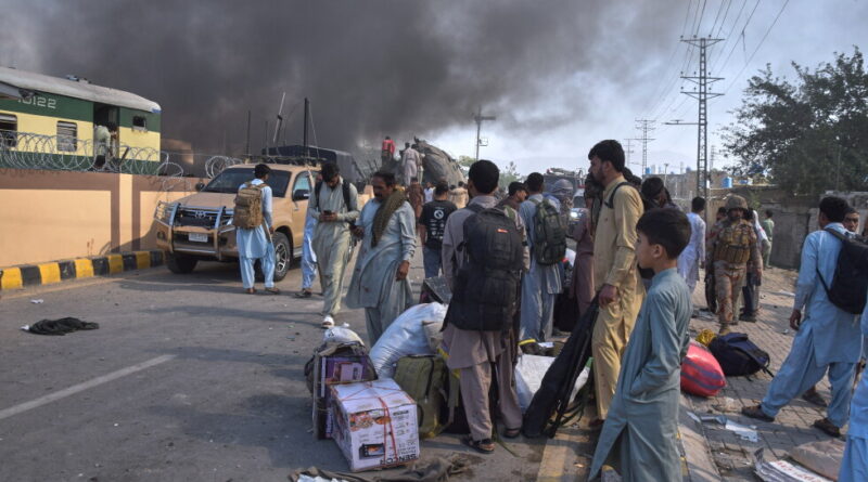Group of civilians with bags and suitcases gathered on a road as a large fire and thick black smoke rise in the distance behind a wrecked vehicle and train car.