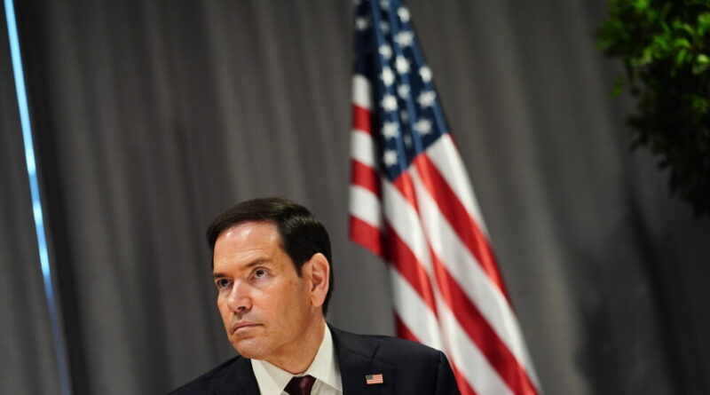 Man in a dark suit and red tie standing in front of an American flag, looking serious.