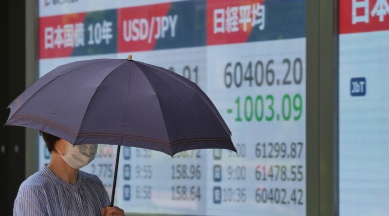 Person wearing a mask holds a dark blue umbrella in front of a large stock market display with numbers and Japanese text in the background.