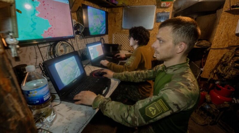 Soldiers in a small, cluttered room monitor weathered maps and digital screens for navigation or briefings at a command workstation.