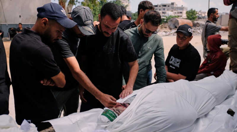 A group of men gathers around a body wrapped in a white shroud outdoors, paying respects near rubble in an open area.