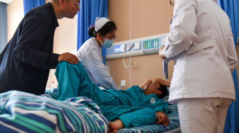 Patient lies in a hospital bed as two nurses and a visitor assist him, with blue curtains in the room.