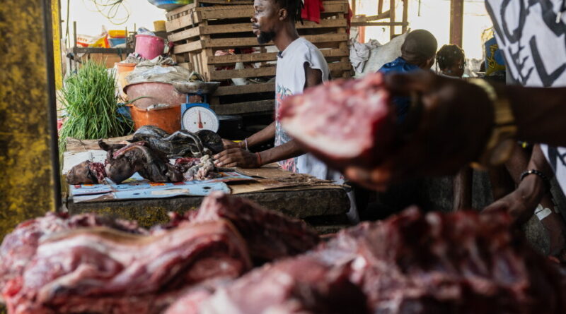 Market butcher at a stall cutting and weighing raw meat with crates and scales in the background.