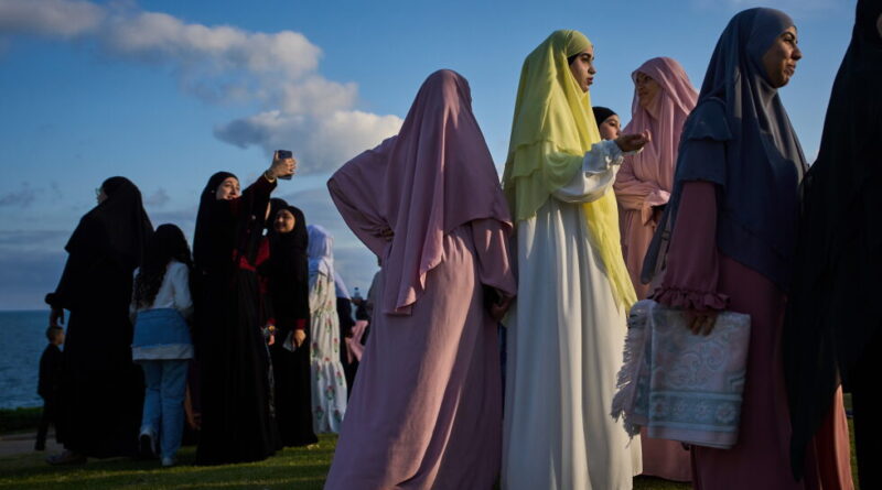 Group of women in pastel hijabs and abayas standing on a grassy coastal area, some chatting and posing by the sea.