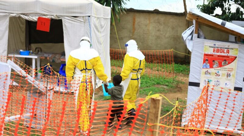 Two health workers in yellow hazmat suits guide a child through an orange safety fence toward a medical tent labeled T1 in a field setting.