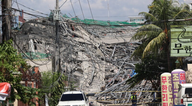 Collapsed building with tangled power lines and rubble, a white SUV in the street, and workers in safety vests securing a cordoned disaster site amid palm trees and signs in Korean.