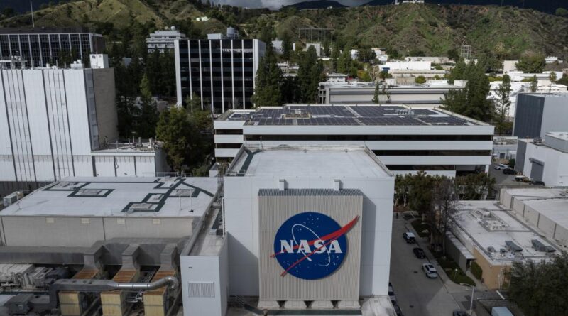 Aerial view of a NASA facility with a large round NASA logo on the building facade, rooftop solar panels visible nearby.