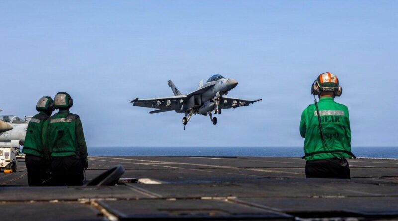 F/A-18 fighter jet landing on an aircraft carrier deck as three crew members in green vests watch from the foreground by the runway edge.