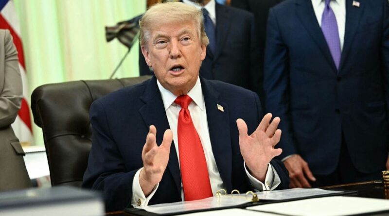 Man in a navy suit with a red tie sits at a desk, gesturing with hands while speaking, with others standing behind him in a formal setting, likely a meeting room.