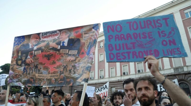 Protesters hold a bright blue placard that reads 'NO TOURIST PARADISE IS ON DESTROYED LIVES' at a street rally, with people and banners in the background.