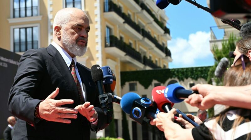 Man in a dark suit giving a press conference outdoors, speaking to multiple microphones held by reporters to his right.