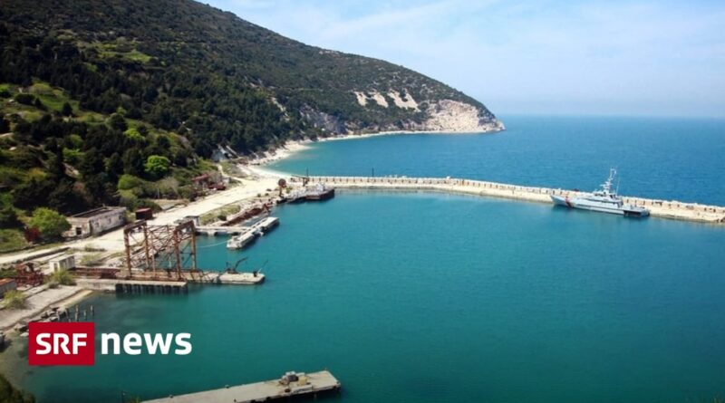 Coastal harbor with a curved breakwater, turquoise water, and a forested hillside; SRF News logo in the bottom-left.