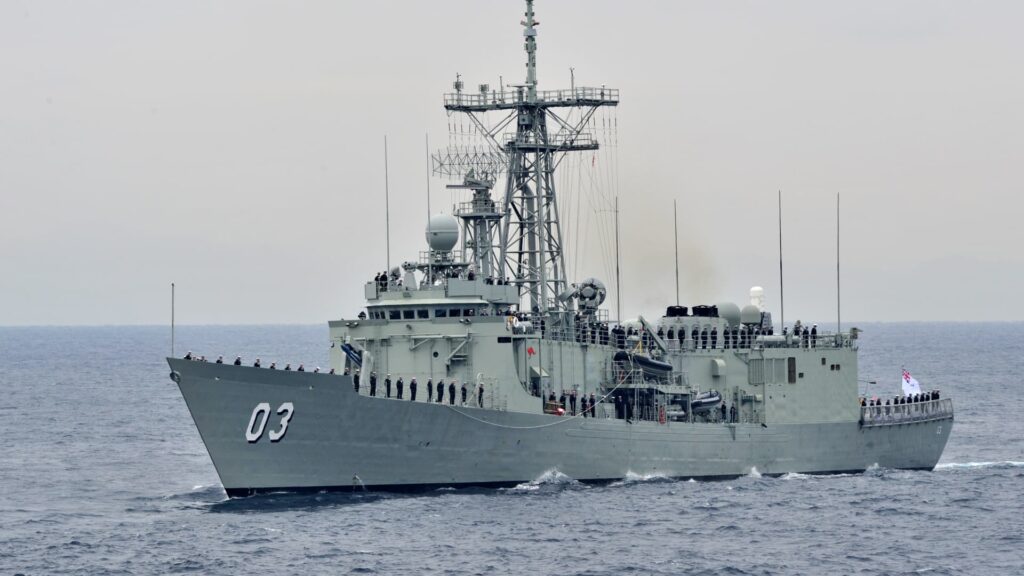 Gray military warship with sailors standing along the railing, sailing on calm sea with a large radar tower ahead.