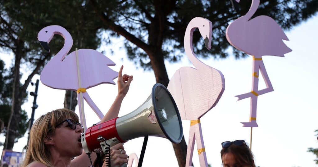Woman wearing sunglasses shouts into a red megaphone at an outdoor event, with tall pink flamingo decorations in the background.