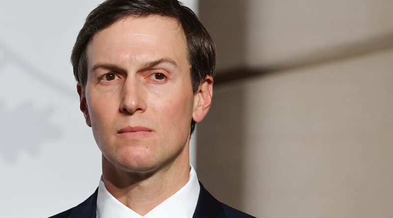 Close-up portrait of a serious-looking man in a dark suit and white shirt, facing slightly left against a neutral background.