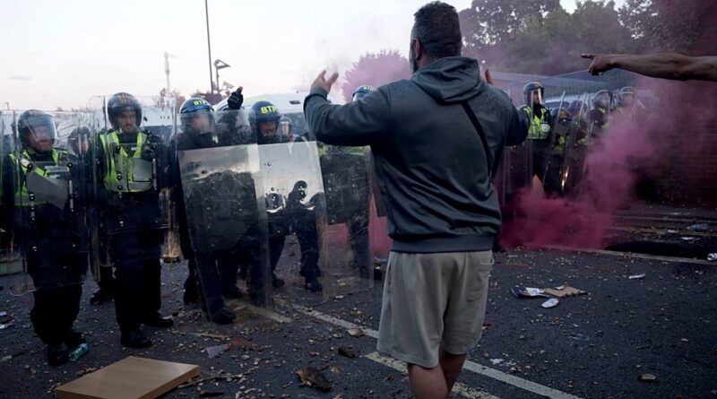 Protester facing a line of riot police with clear shields, pink smoke in the air, street debris scattered around.