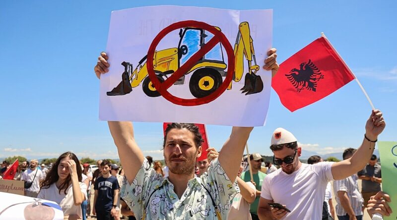 Protesters hold signs outdoors; the central poster shows a construction excavator with a red prohibited symbol, with an Albanian flag in the background.