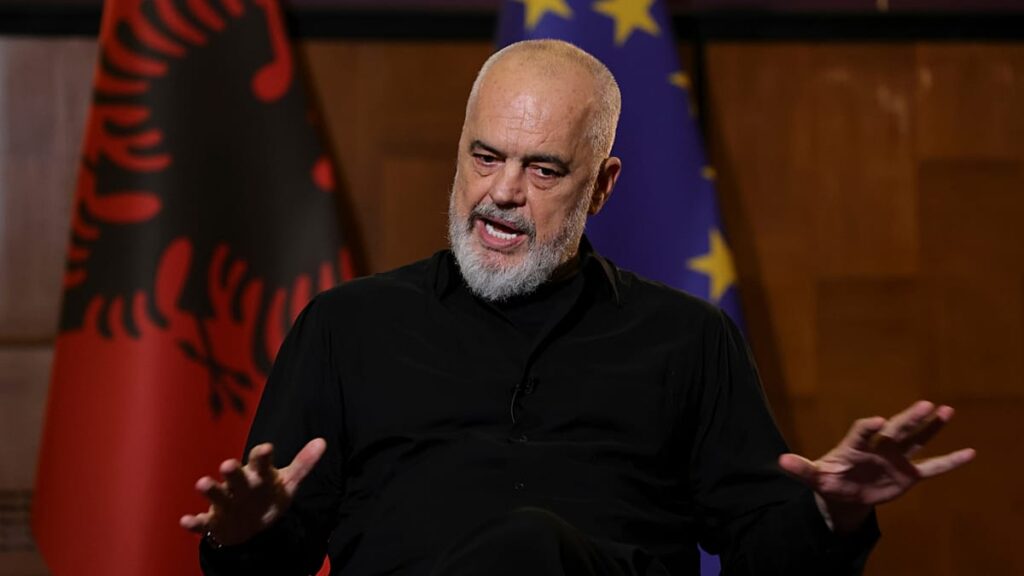 Bearded man in a black shirt speaks during an interview, with Albanian and EU flags visible behind him.