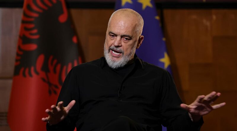 Bearded man in a black shirt speaks during an interview, with Albanian and EU flags visible behind him.