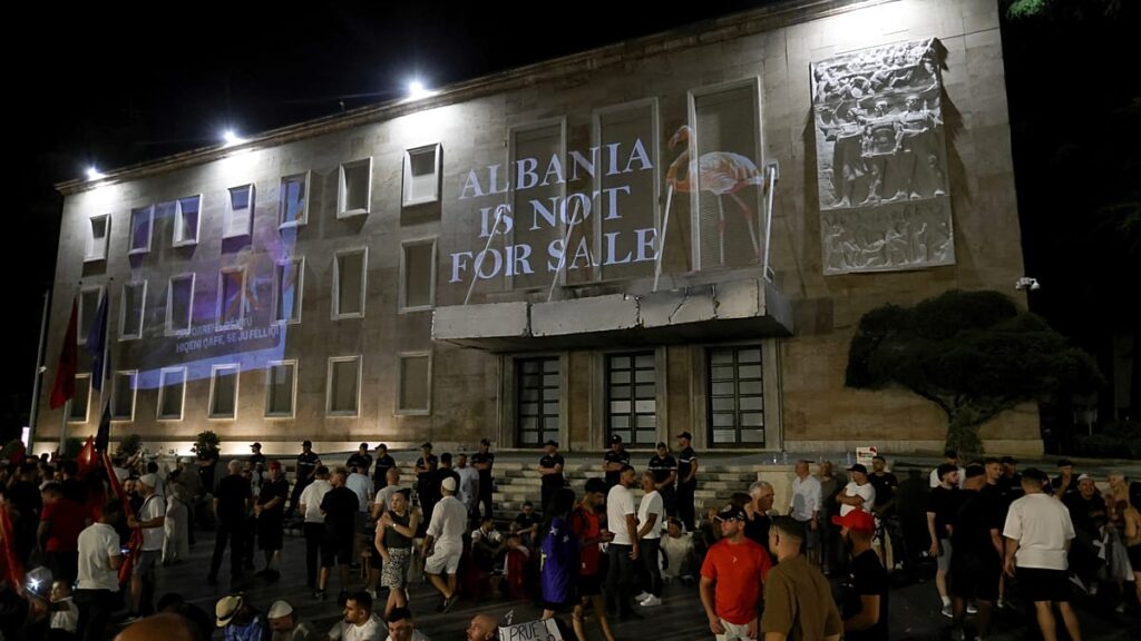 Night scene outside a large stone building with a prominent banner reading 'ALBANIA IS NOT FOR SALE' and a crowd gathered in front, some people seated on steps.