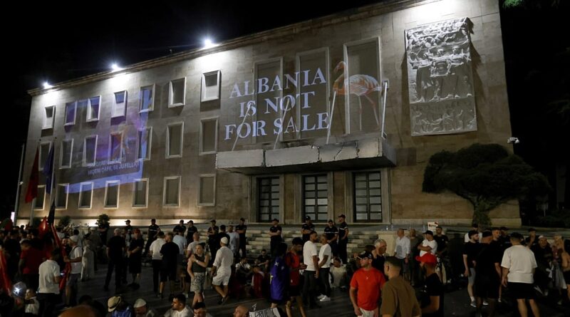 Night scene outside a large stone building with a prominent banner reading 'ALBANIA IS NOT FOR SALE' and a crowd gathered in front, some people seated on steps.