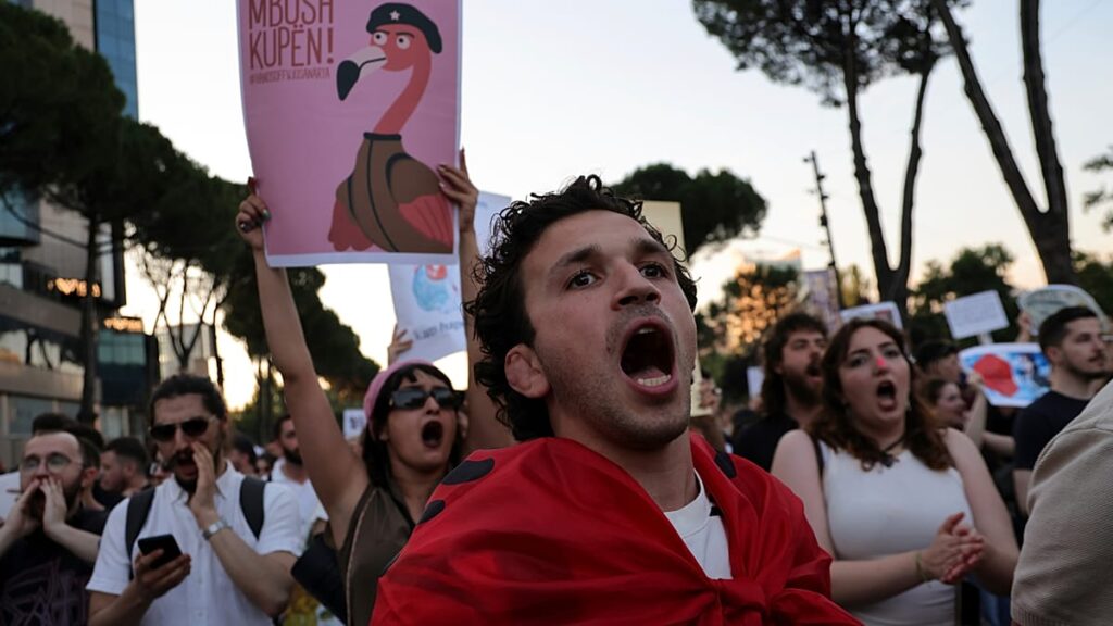 Young man in a red scarf shouts at a protest, surrounded by chanting protesters with signs at dusk.