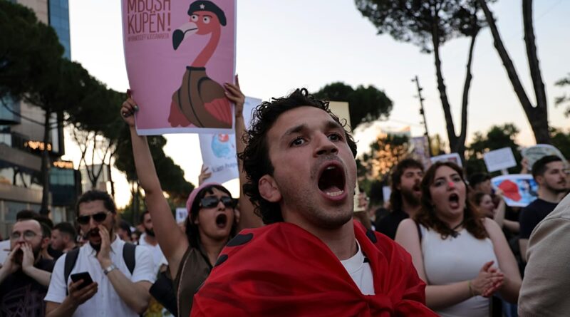 Young man in a red scarf shouts at a protest, surrounded by chanting protesters with signs at dusk.