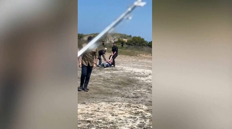Group of people helping an injured person lying on the ground in an open, rocky area; a diagonal pole crosses the frame.