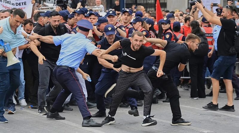 Crowd of police and protesters clash in a street confrontation; a man in a black shirt lunges forward while others pull him.