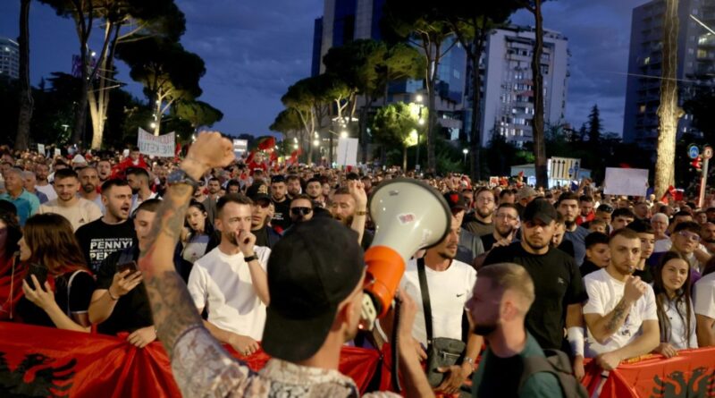 Large crowd of protesters at night with a speaker using a megaphone at the front, raising a fist and banners in the background.