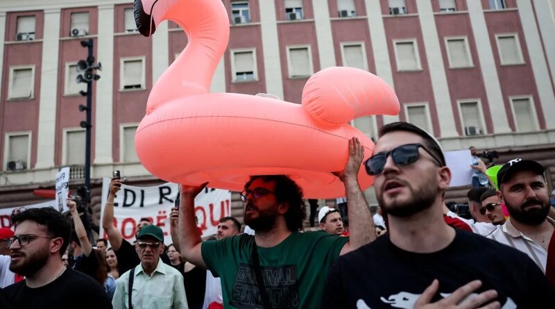 Crowd of protesters in an urban square, a man holds a large pink inflatable flamingo above his head.