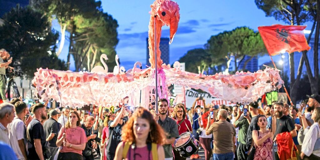 Crowd at a street parade beneath a giant pink paper flamingo banner.