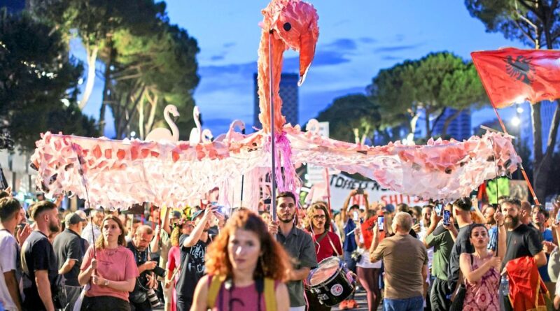 Crowd at a street parade beneath a giant pink paper flamingo banner.